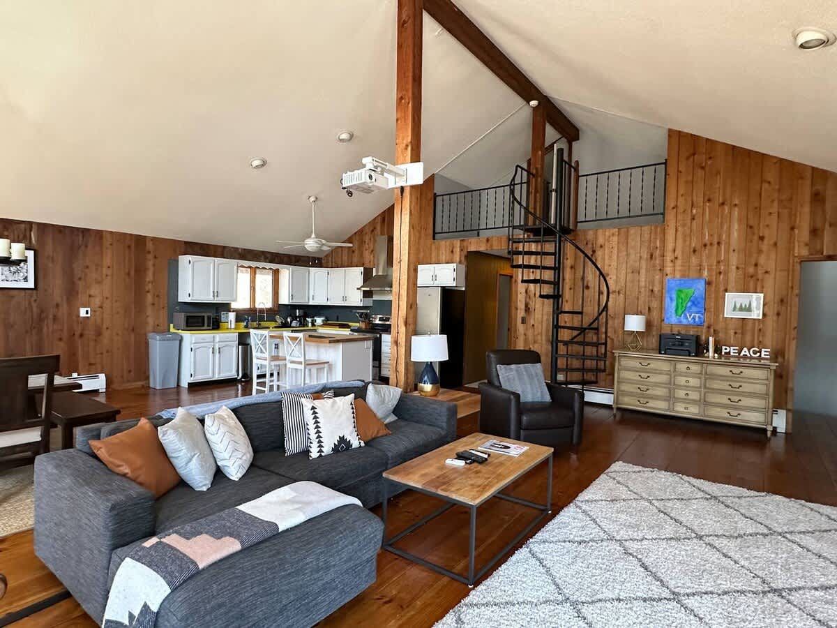 Spacious open-concept living area flowing into the kitchen, featuring a large grey sectional sofa, a leather armchair, and a black spiral staircase, all under a vaulted ceiling with exposed wood beams.