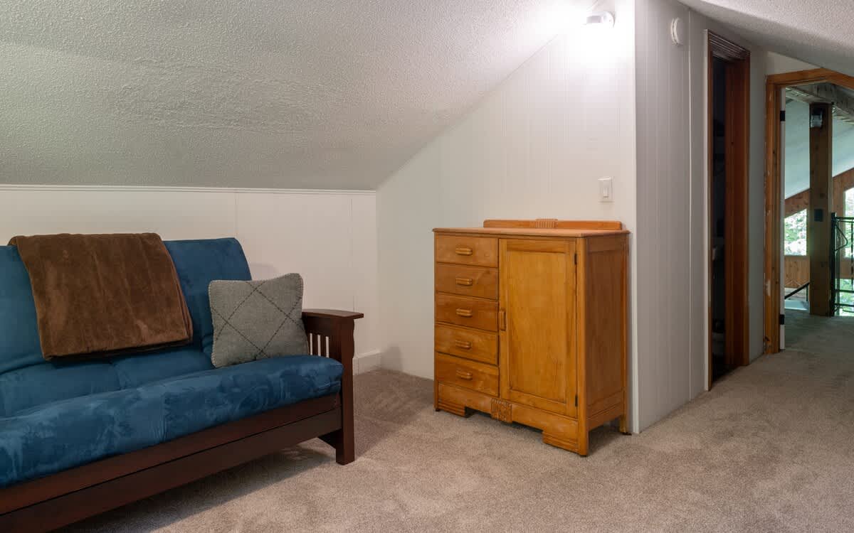 Carpeted loft area with a blue futon, a wooden dresser, and white paneled walls with a sloped ceiling, offering a glimpse into an adjacent hallway.