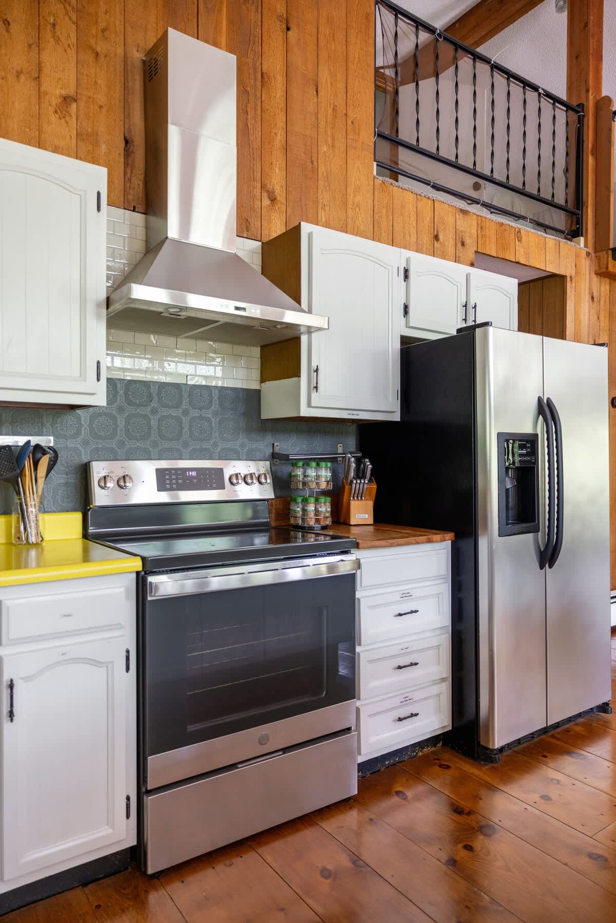 Modern kitchen area showcasing a stainless steel electric range with matching hood, a large stainless steel French door refrigerator, and white cabinets with wood countertops. A gray patterned backsplash adds texture.