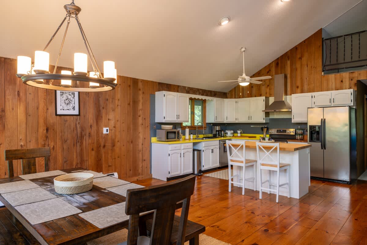 Open-concept dining area adjacent to the kitchen, featuring a rustic wooden dining table with benches and chairs, illuminated by a wagon-wheel style chandelier. Wood-paneled walls and hardwood floors complete the look.