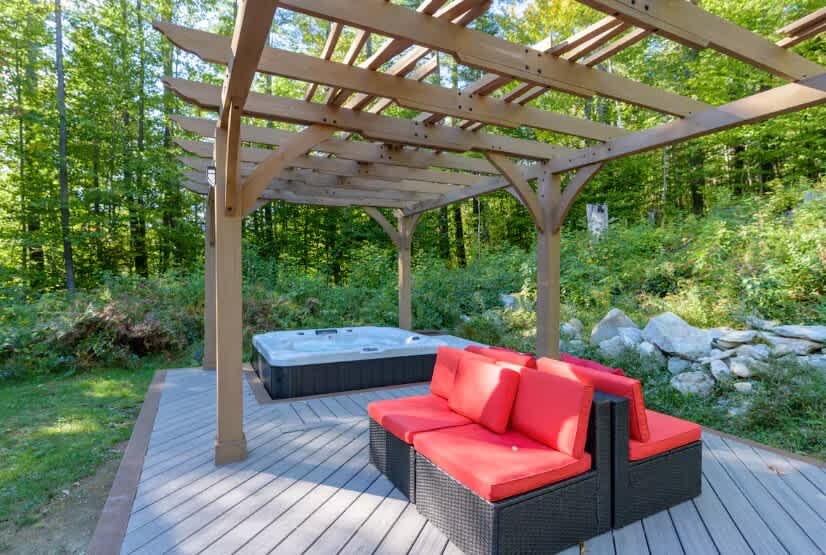 Outdoor relaxation area featuring a hot tub and black wicker seating with vibrant red cushions, all situated on a wooden deck under a rustic wooden pergola, surrounded by lush greenery.
