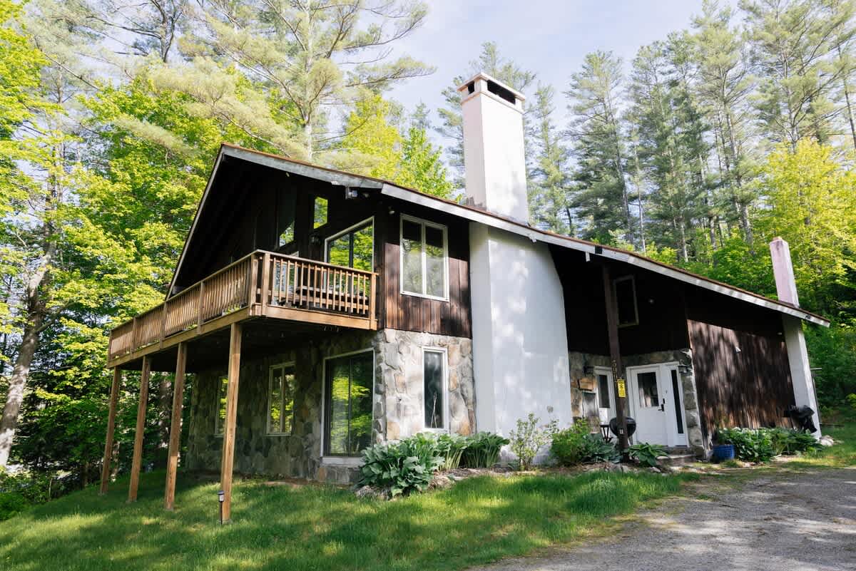 Front view of a rustic chalet-style house featuring a red metal roof, dark wood and stone siding, a prominent white chimney, and a wooden balcony, set amidst a green forest.