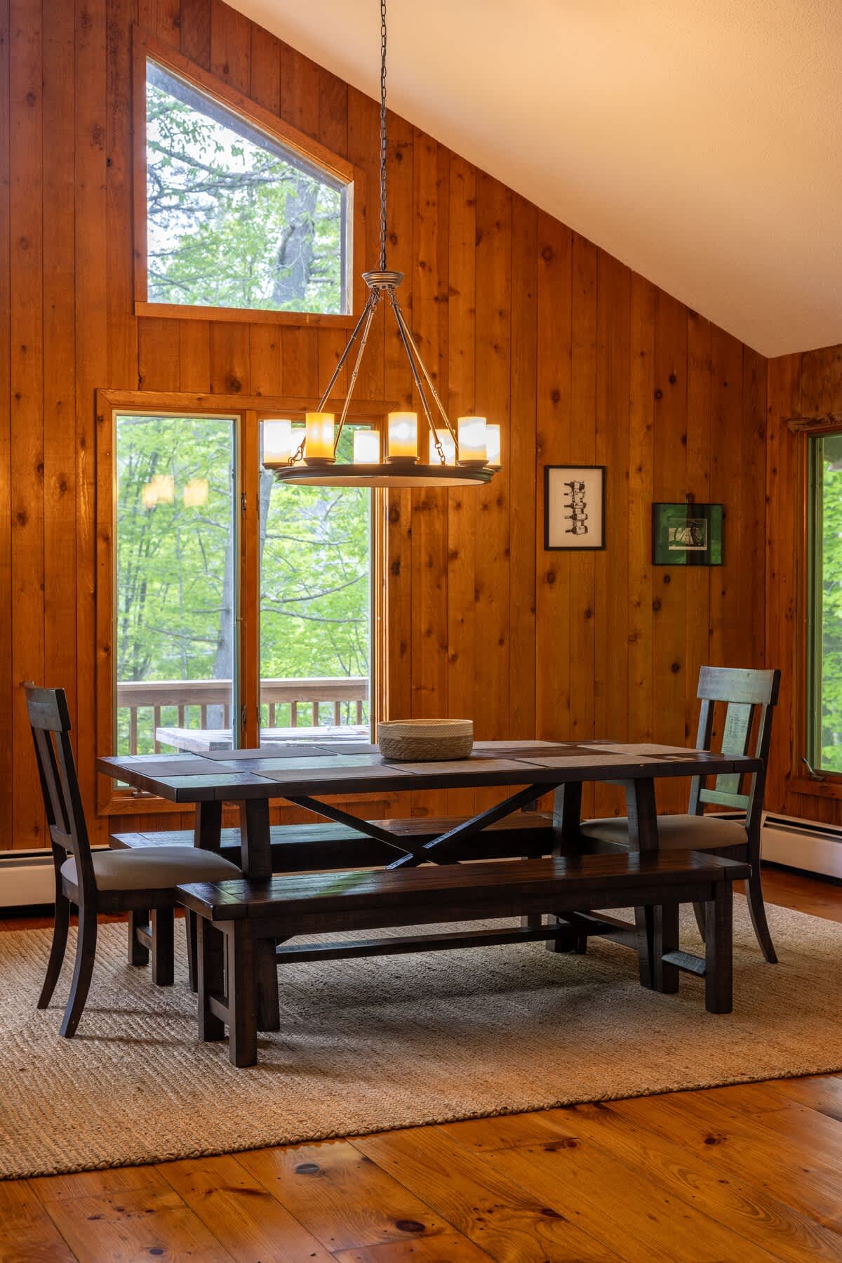 Inviting dining area with a rustic wooden table and bench seating, set against a backdrop of large windows that provide ample natural light and views of lush greenery. A chandelier hangs above the table.