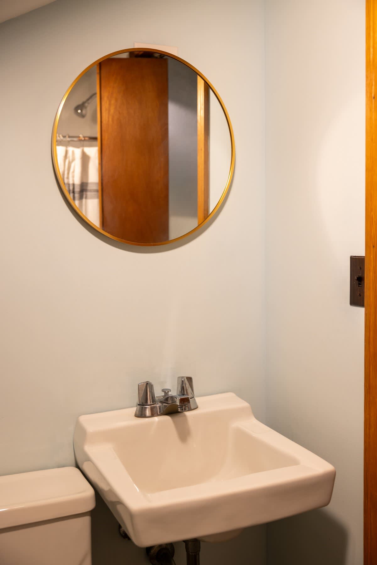 A compact bathroom featuring light blue walls, a white ceramic sink with chrome faucets, and a round gold-framed mirror reflecting a shower area with a striped curtain and a wooden door.