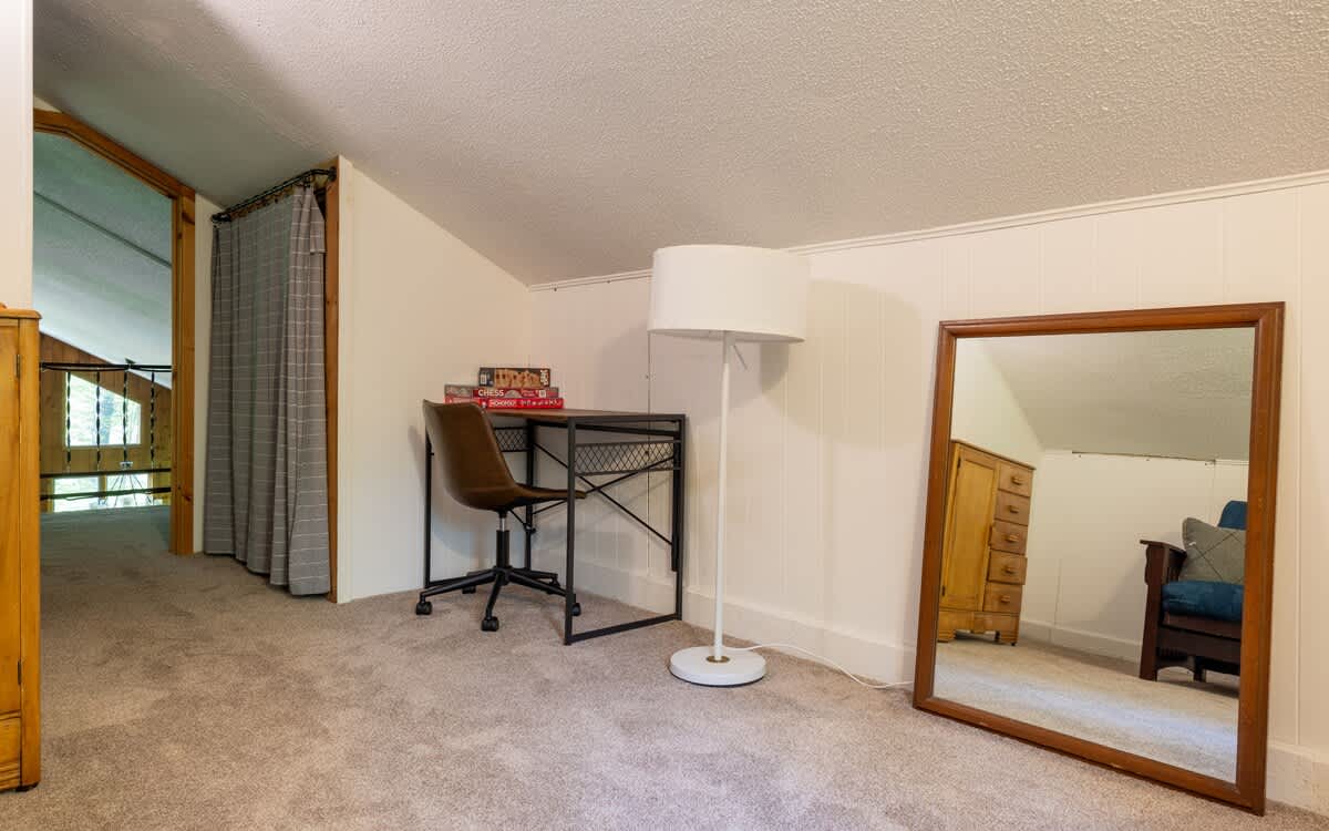 Carpeted loft area with a desk, office chair, floor lamp, and a large full-length mirror, featuring white paneled walls and a sloped ceiling.