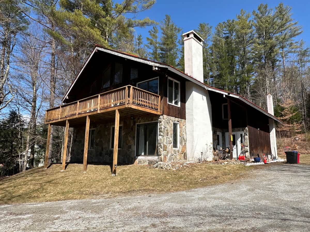 A-frame style house with a mix of stone facade and dark wood siding, featuring a large wooden deck on the upper level. Surrounded by tall pine trees under a clear blue sky, with a gravel driveway.