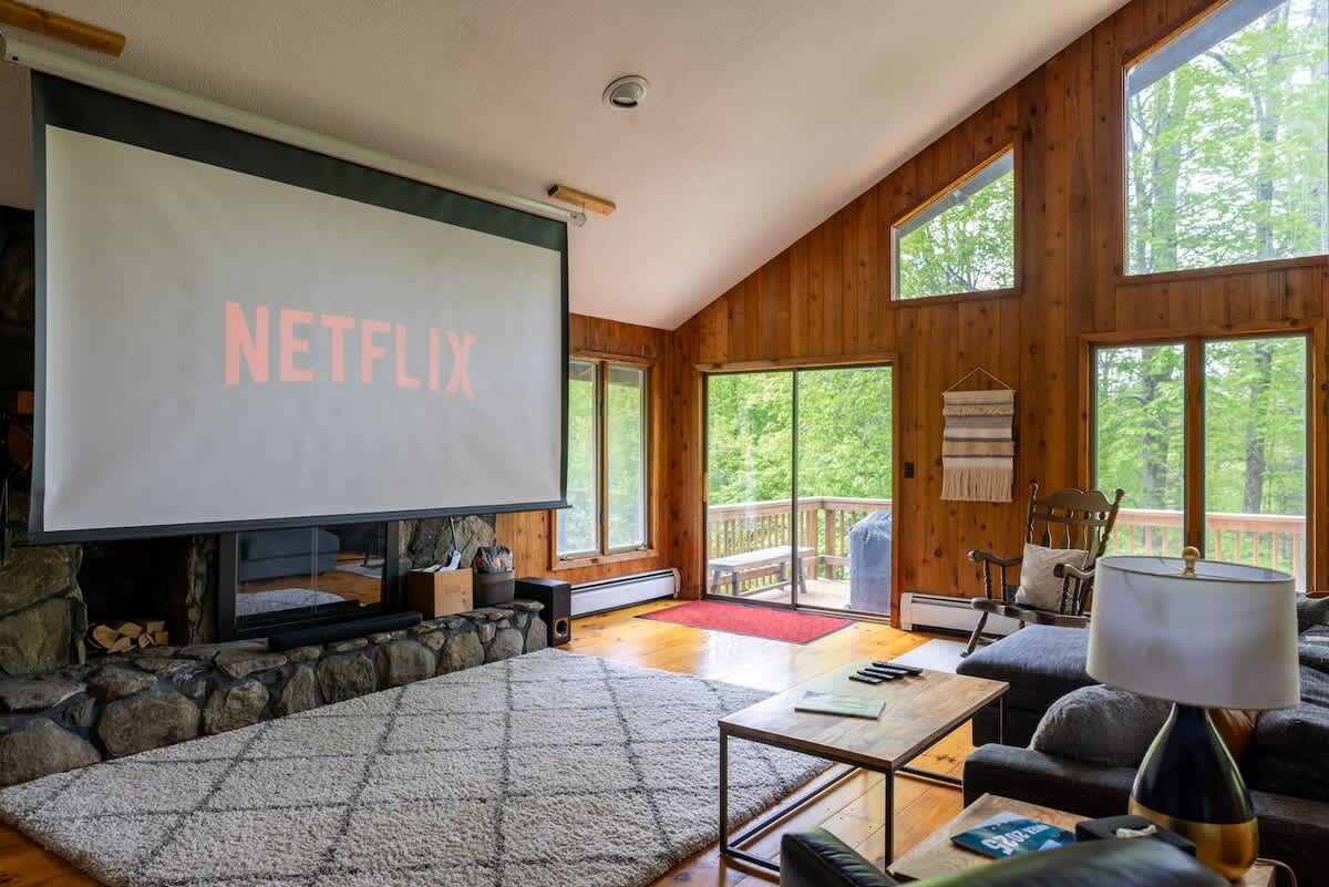 Entertainment-focused living space with a large projector screen displaying 'NETFLIX' above a stone fireplace, surrounded by expansive windows offering views of the surrounding lush trees.