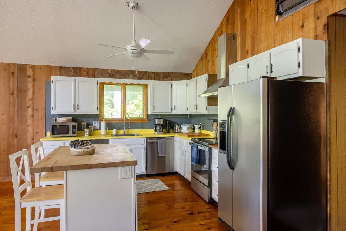 Bright kitchen featuring white shaker cabinets, vibrant yellow countertops, and stainless steel appliances including a refrigerator, dishwasher, and microwave. Wood-paneled walls and hardwood floors add a rustic touch.