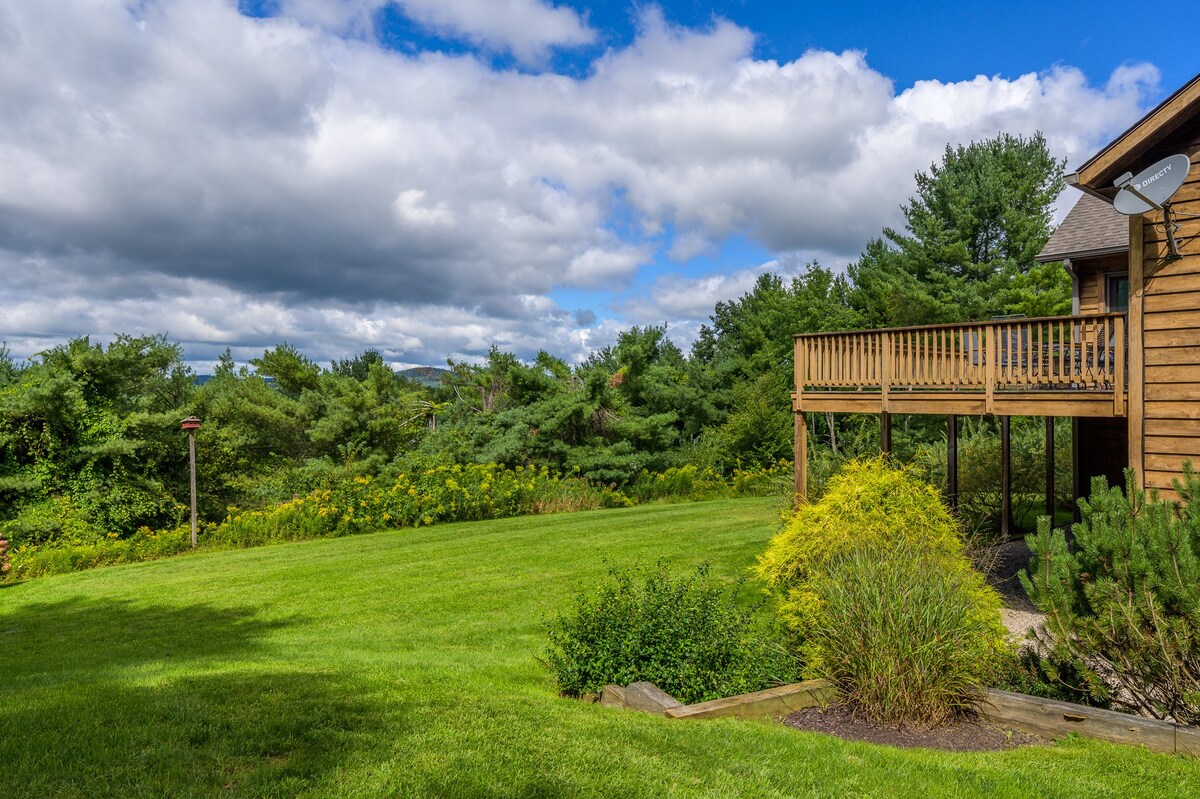 A sprawling green lawn slopes gently down towards a dense forest, offering sweeping views of distant hills under a partly cloudy sky. A wooden deck with a railing extends from the side of a rustic wooden house, overlooking the scenic landscape.