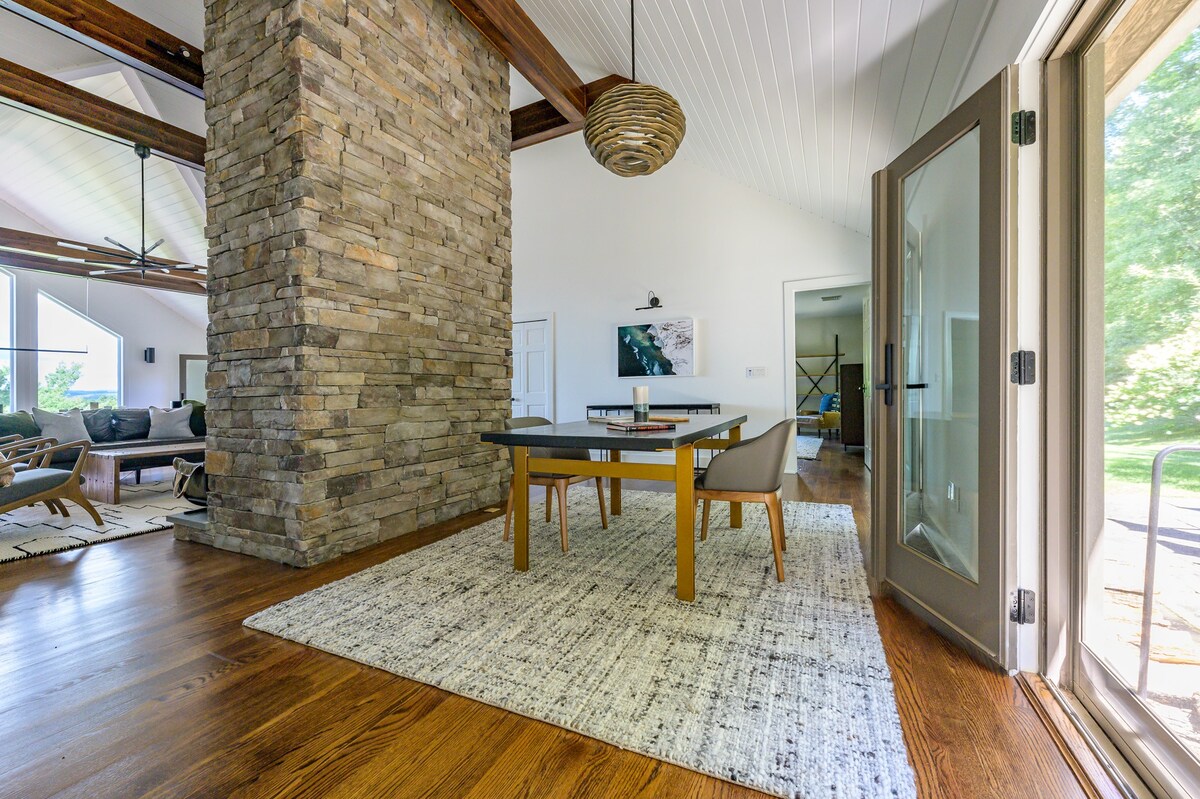 An inviting entry area features a prominent stone column, a small dining or game table with two chairs on a patterned rug, and hardwood floors, with a glimpse of the living room beyond and an open door to the outside.