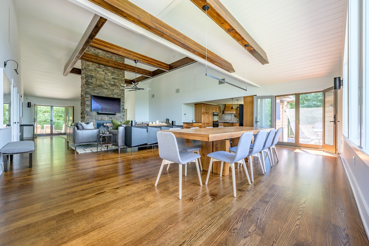 A spacious open-plan area showcases a long wooden dining table with light grey chairs, leading into the living room with a stone fireplace and leather sofas, all featuring hardwood floors and vaulted ceilings with exposed beams.