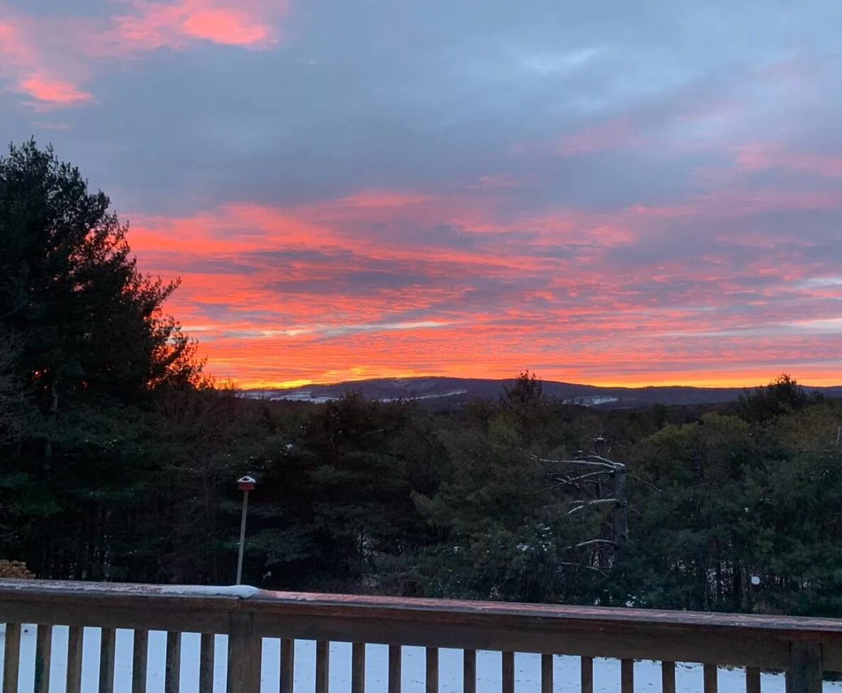 A breathtaking sunset paints the sky with vibrant reds and oranges over a snow-covered landscape and dense forest. The view is framed by a wooden deck railing in the foreground, highlighting the expansive natural beauty.