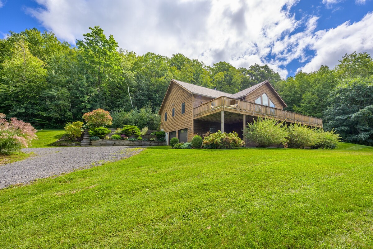A rustic wooden house with a gabled roof is nestled on a hillside, surrounded by lush green trees and a well-maintained lawn. A gravel driveway leads up to the house, which features a large elevated deck with a wooden railing.
