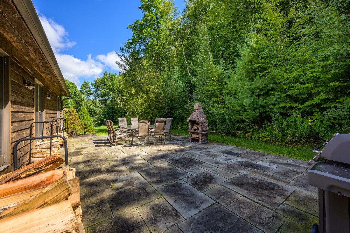 A spacious stone patio is surrounded by dense green trees and features an outdoor dining set and a stone outdoor fireplace. Stacked firewood is neatly arranged against the rustic wooden house, and a BBQ grill is visible on the right.
