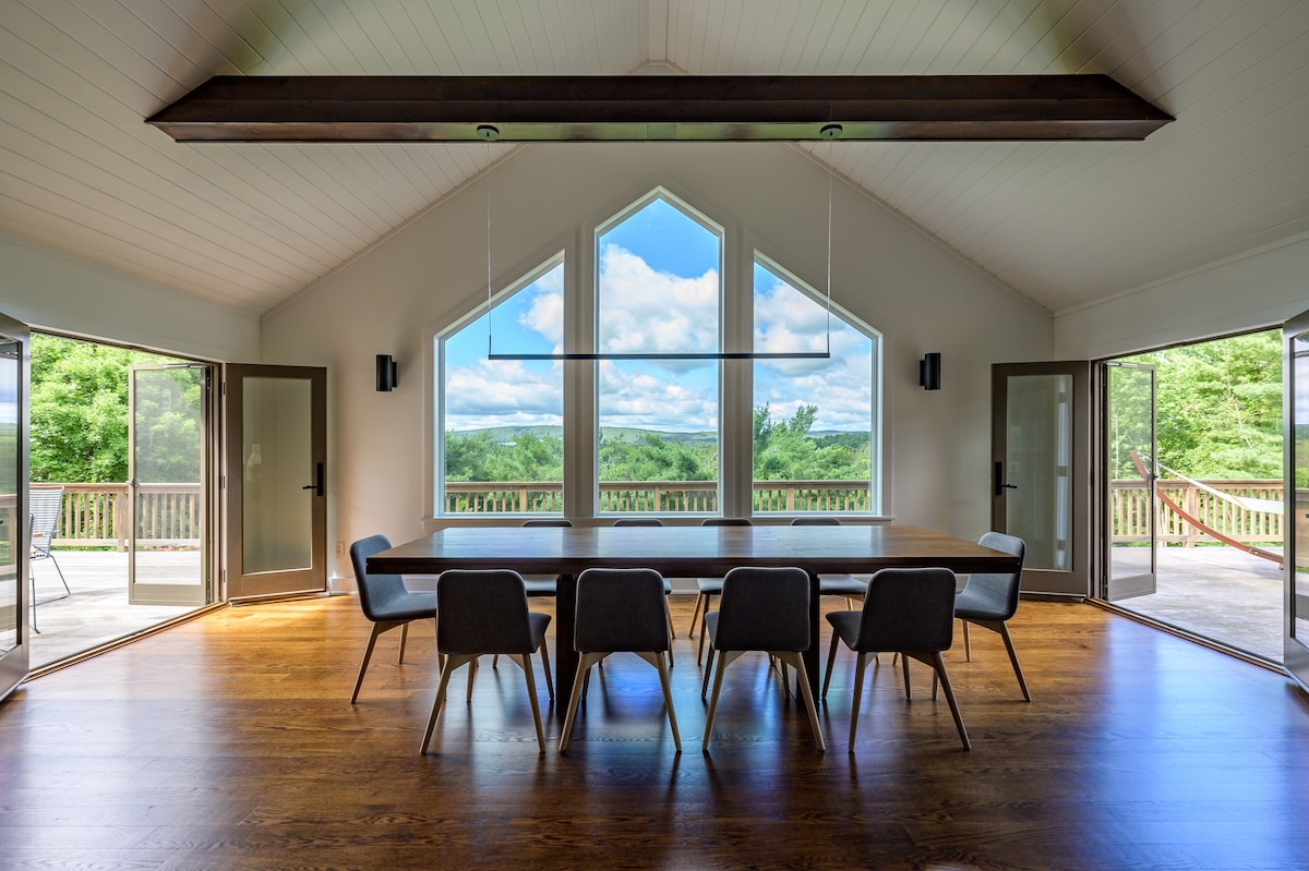 A bright dining room features a long wooden table with modern chairs, positioned to showcase expansive mountain views through large, geometrically shaped windows, complemented by vaulted ceilings and hardwood floors.