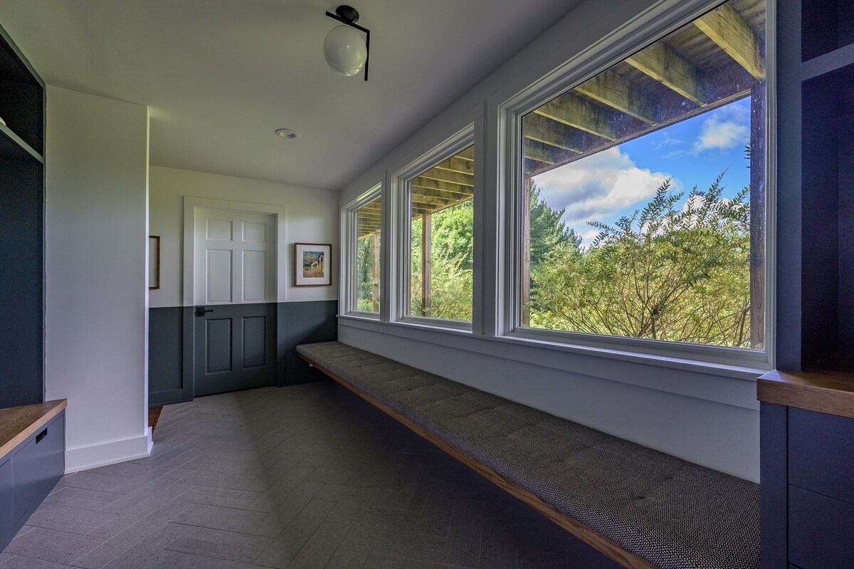 A bright and modern mudroom features a long built-in bench with a cushion, flanked by storage cabinets. Large windows offer a view of lush greenery outside, and the room has light walls with a dark wainscoting and herringbone-patterned floor tiles.