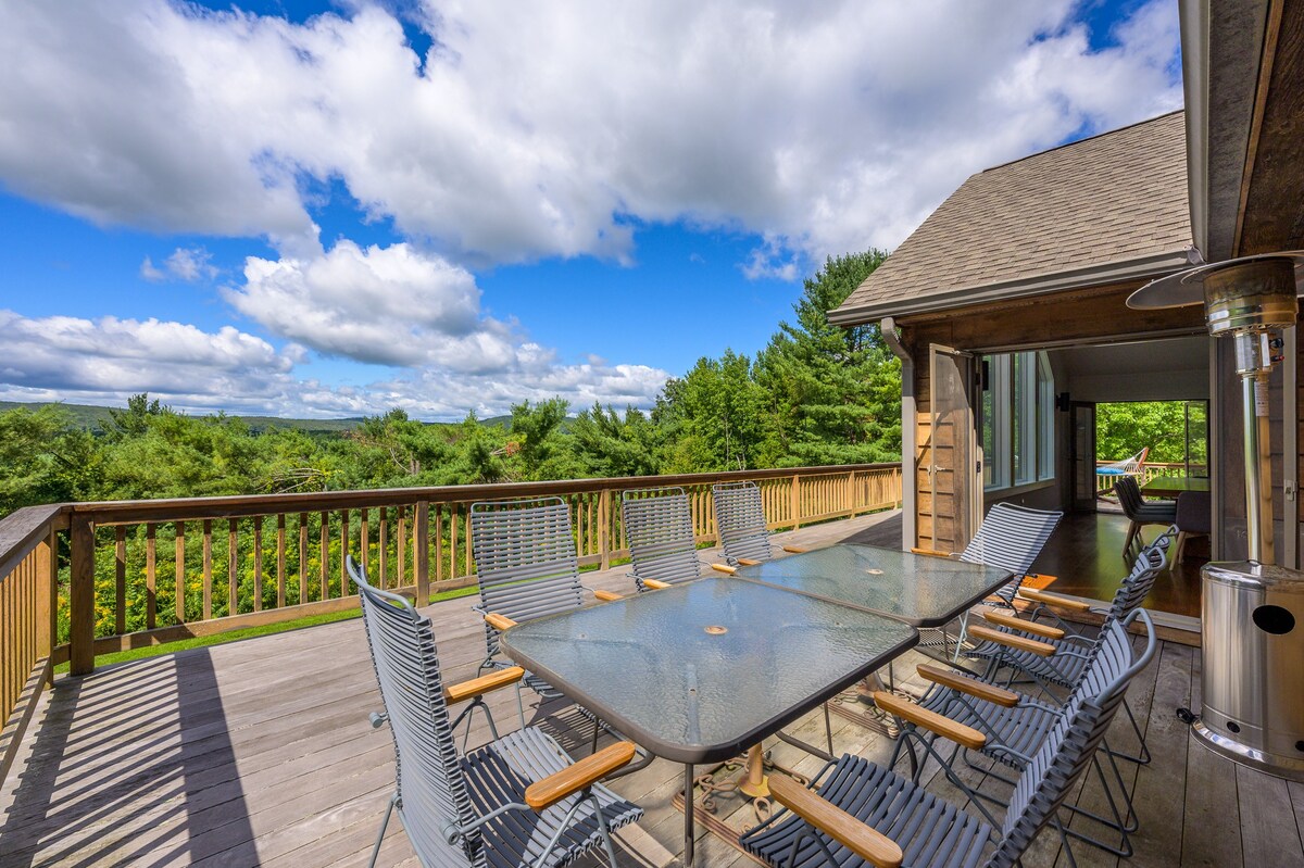 A spacious wooden deck offers an outdoor dining area with multiple tables and chairs, overlooking a vast expanse of green forest and distant hills under a bright blue sky with white clouds.