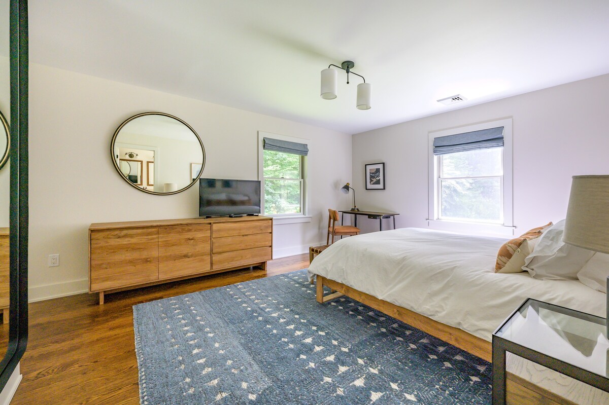 Modern bedroom featuring a wooden bed frame, a long wooden dresser with a TV, and a round gold mirror. A blue geometric rug covers dark hardwood floors, and two windows offer natural light.