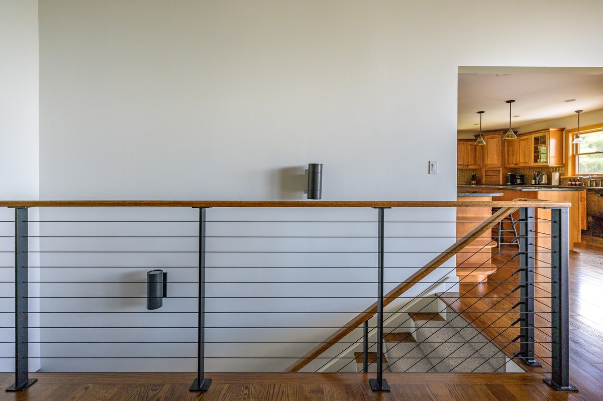 A modern landing area features a cable railing system with a wooden handrail, overlooking a staircase. In the background, a kitchen with light wood cabinetry and pendant lights is visible.