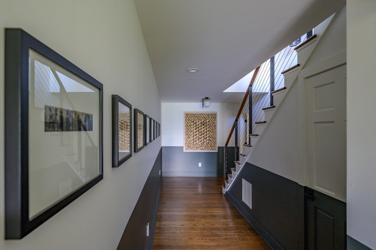 A modern hallway features a two-tone wall with a dark gray wainscoting and light upper section, dark hardwood floors, and a staircase with cable railings. Framed artwork lines one wall, and a textured art piece hangs at the end of the hall.