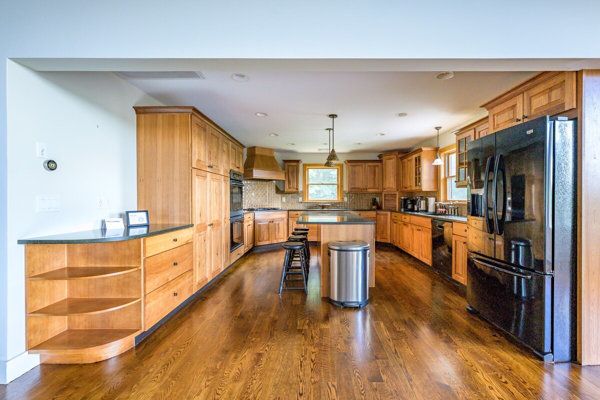 A wide shot of a large kitchen with light wood cabinetry wrapping around the perimeter, dark countertops, and a central island with a stainless steel trash can. Black appliances, including a refrigerator and oven, are integrated into the design, and hardwood floors extend throughout.