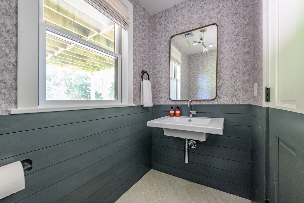 A stylish powder room features a dark gray shiplap wainscoting and patterned wallpaper above, with a white floating sink and a rectangular mirror. A window provides natural light, and the floor has a light-colored herringbone tile pattern.