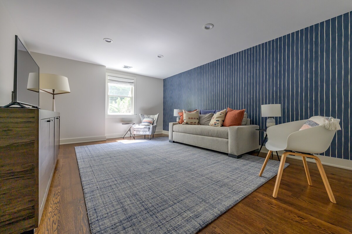 A spacious living room features a light gray sofa with colorful throw pillows, a large blue patterned rug over dark hardwood floors, and a striking navy blue accent wall with white vertical stripes. A flat-screen TV is visible on a wooden console.