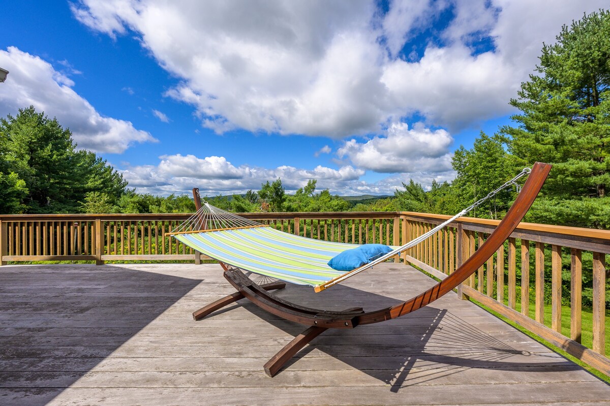 A wooden deck features a comfortable hammock with a blue pillow, offering a serene spot to relax while enjoying the sunny weather and panoramic views of the surrounding green trees and distant landscape.