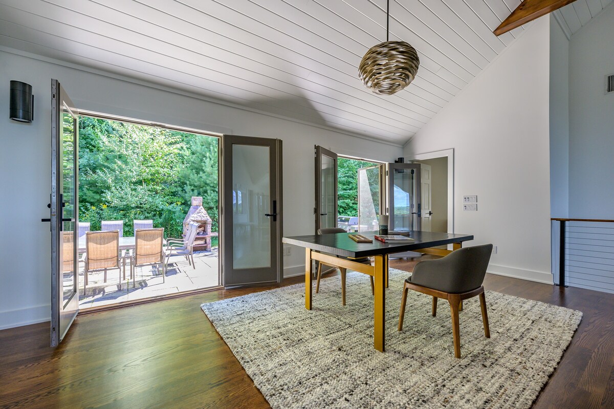A bright room with vaulted ceilings and dark hardwood floors features a modern black table with a light wood base and gray chairs, set on a textured area rug. French doors open to an outdoor patio with seating and lush greenery.