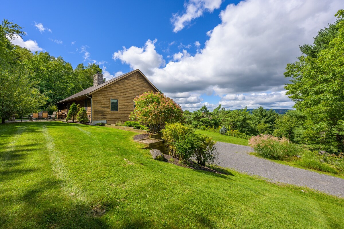 A rustic wooden house with a stone chimney is situated on a gently sloping green lawn, surrounded by mature trees and landscaped bushes. A gravel path is visible on the right, leading towards the house, which offers views of the distant landscape.