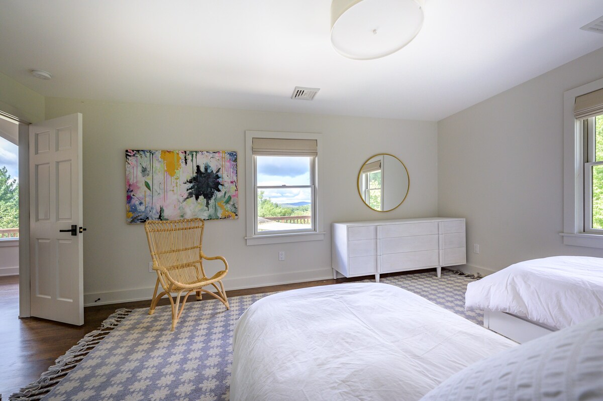 Bright bedroom with light gray walls, two white-sheeted beds, a white dresser, a round gold mirror, and a colorful abstract painting. A rattan chair sits on a blue and white geometric rug, with a large window offering outdoor views.