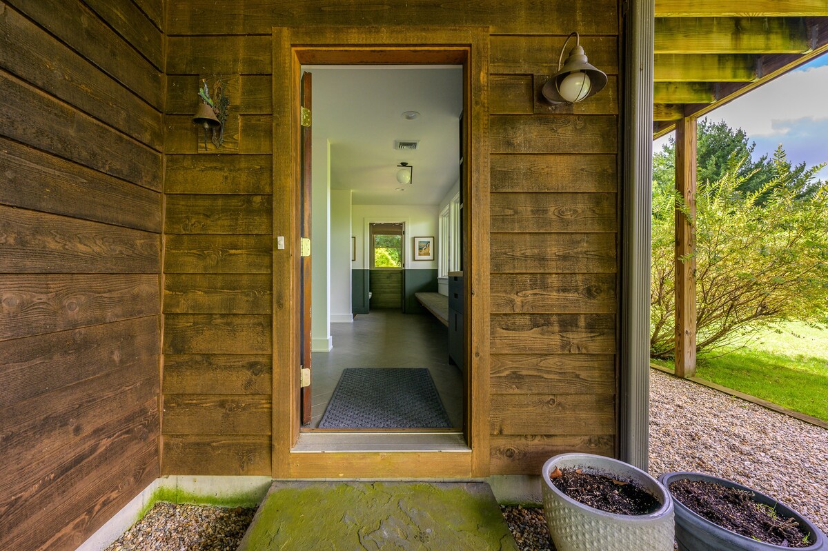 The main entrance features a rustic wooden door set into a wood-paneled exterior, leading into a bright interior hallway. A light fixture hangs above the door, and potted plants are visible outside.