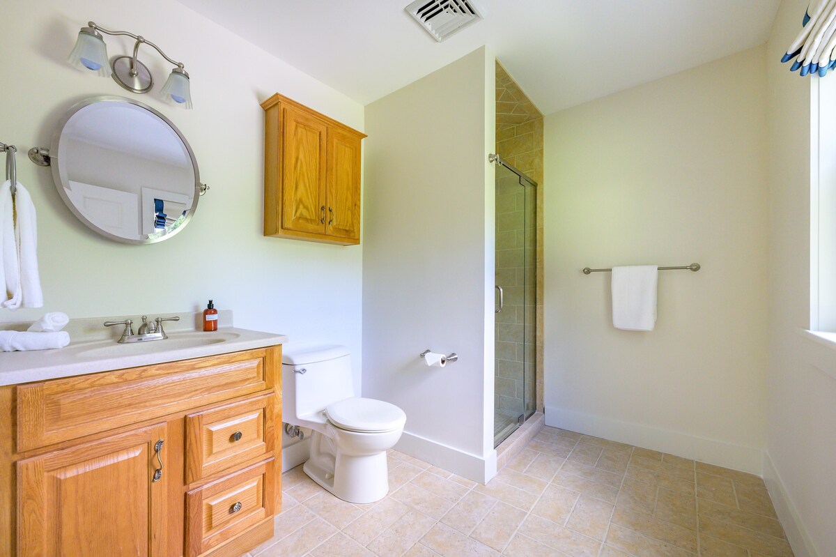 Bright bathroom with a wooden single vanity, an oval mirror, and a toilet. A walk-in shower with light brown tiled walls is visible, and the floor is covered in matching tiles.