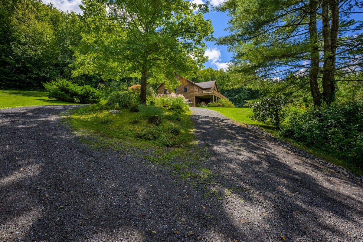 A gravel driveway leads up to a multi-level wooden chalet nestled among vibrant green trees and manicured lawns. The house features multiple windows and a visible deck, all under a bright sky.