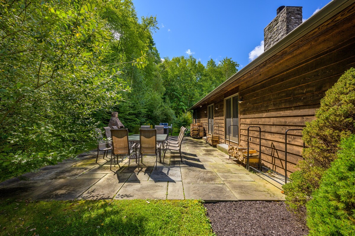 A stone patio is set amidst lush green trees and foliage, featuring an outdoor dining table with chairs and a stone outdoor fireplace. The side of a rustic wooden house with a stone chimney is visible, along with stacked firewood.