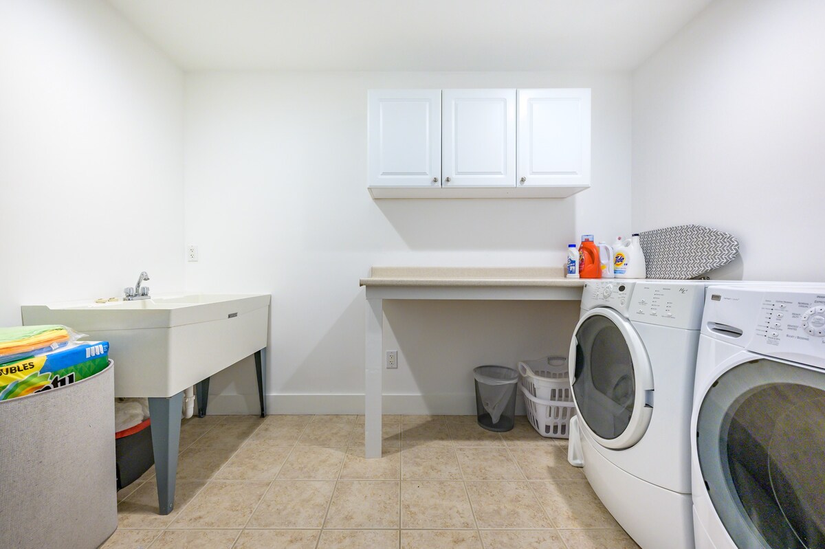 Functional laundry room with a white utility sink, a front-loading washer and dryer, and white wall cabinets. A long countertop provides space for folding, all set against light tiled floors.