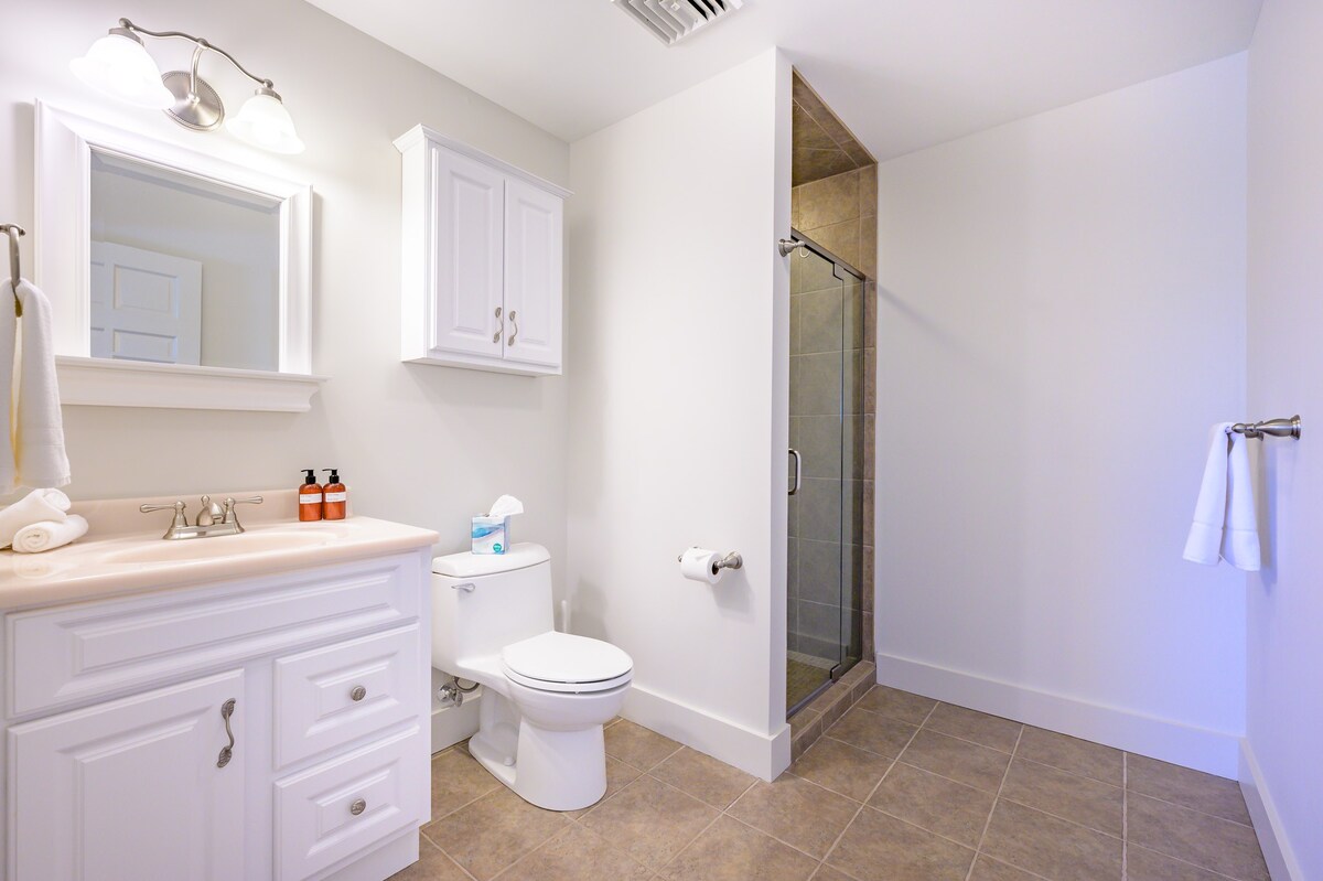 Clean white bathroom featuring a single vanity with a light pink countertop and white shaker-style cabinets. It includes a toilet and a walk-in shower with brown tiled walls and a glass door.