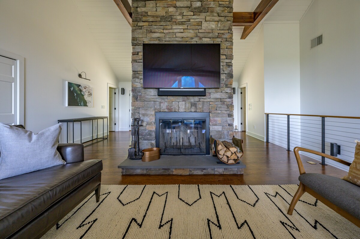 A detailed view of the living room's impressive floor-to-ceiling stone fireplace, featuring a mounted television and a burning fire, surrounded by a patterned rug and dark hardwood floors, with a glimpse of a leather sofa.