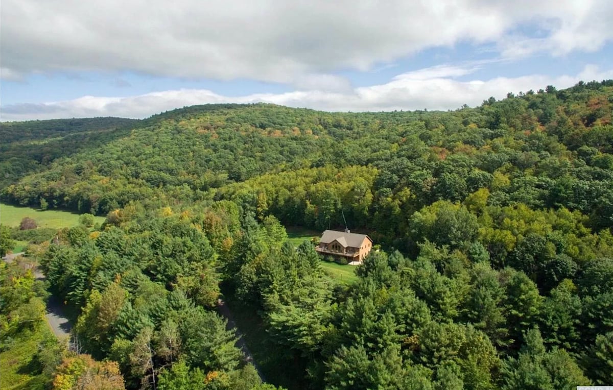 An aerial view captures the chalet-style house nestled amidst a dense, verdant forest, with rolling hills and mountains in the distance under a partly cloudy sky, highlighting its secluded natural setting.