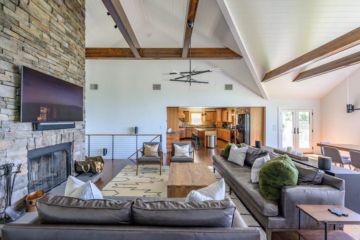 An open-concept living space features a stone fireplace as a central element, with a large leather sectional and a wooden coffee table, leading into a visible kitchen area in the background, all under vaulted ceilings with exposed wooden beams.