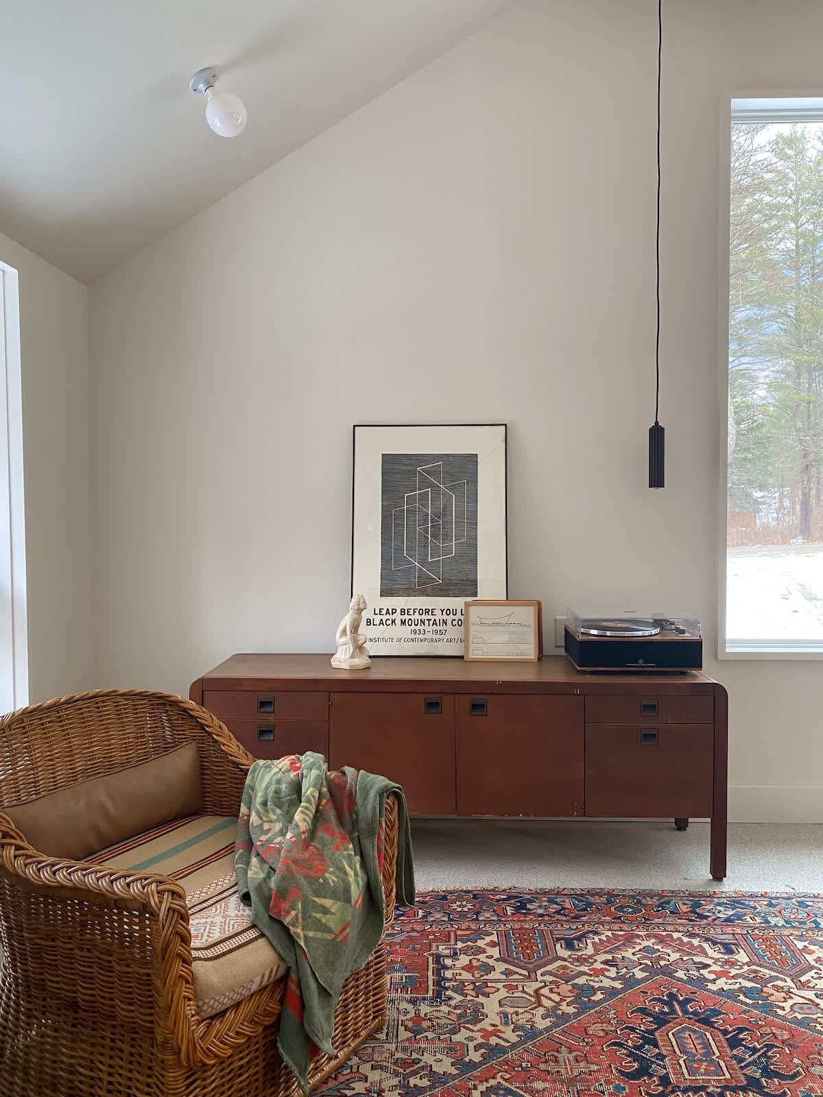 A close-up of a living room corner featuring a wooden credenza with a record player and framed artwork, next to a wicker armchair with a patterned blanket, all on a colorful area rug.