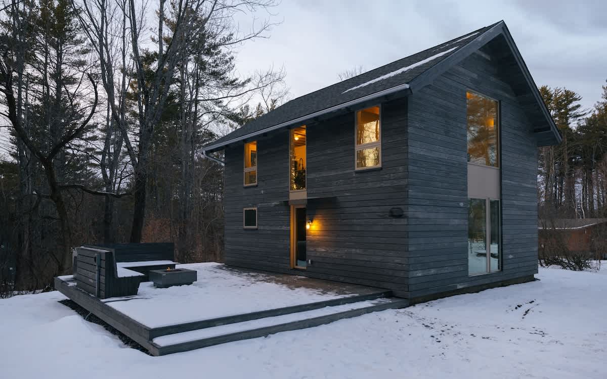 The modern dark grey house stands against a twilight sky, with warm light emanating from its windows, illuminating the snowy ground and the adjacent deck with a fire pit.