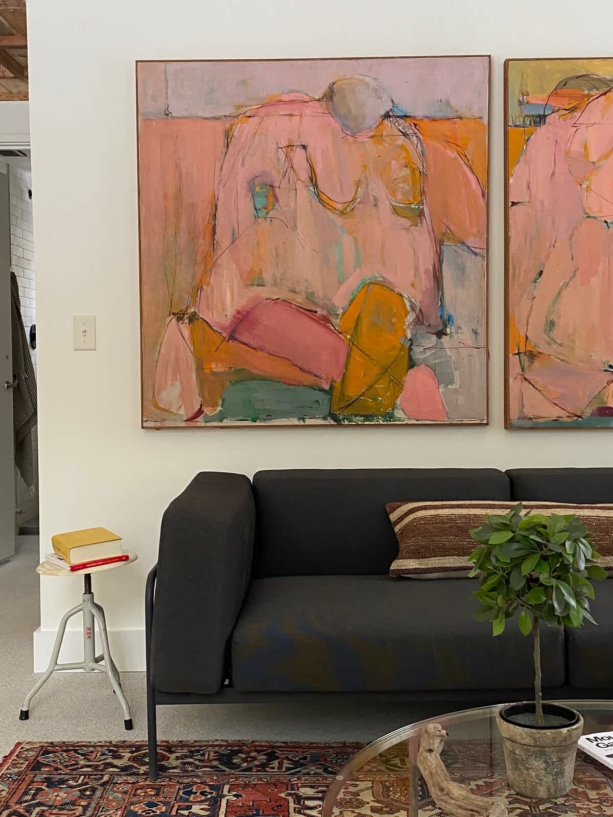 A cozy living room features a dark grey sofa with a striped pillow, a glass-top coffee table with a small potted plant, and a colorful abstract painting dominating the white wall above. A patterned rug covers the floor.