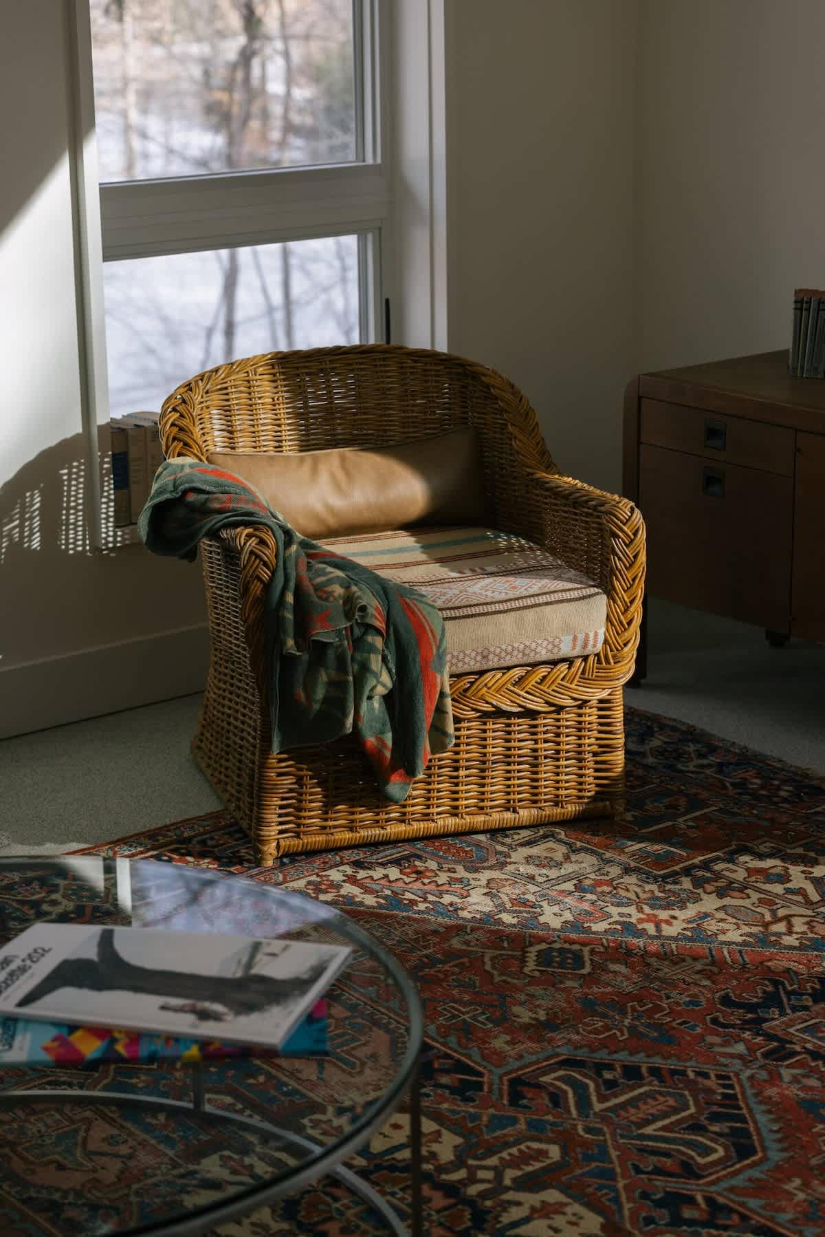 A cozy corner of a living space featuring a wicker armchair with a patterned cushion and a green and orange throw blanket. A vintage-style rug with intricate patterns covers the floor, and a round glass-top coffee table holds magazines.