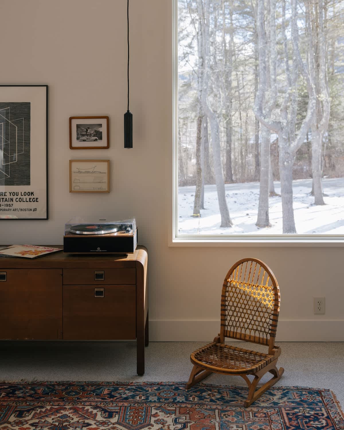 A close-up of the living area shows a wooden sideboard with a record player and framed art, next to a large window revealing a snow-covered landscape and bare trees. A woven chair sits on a patterned rug.