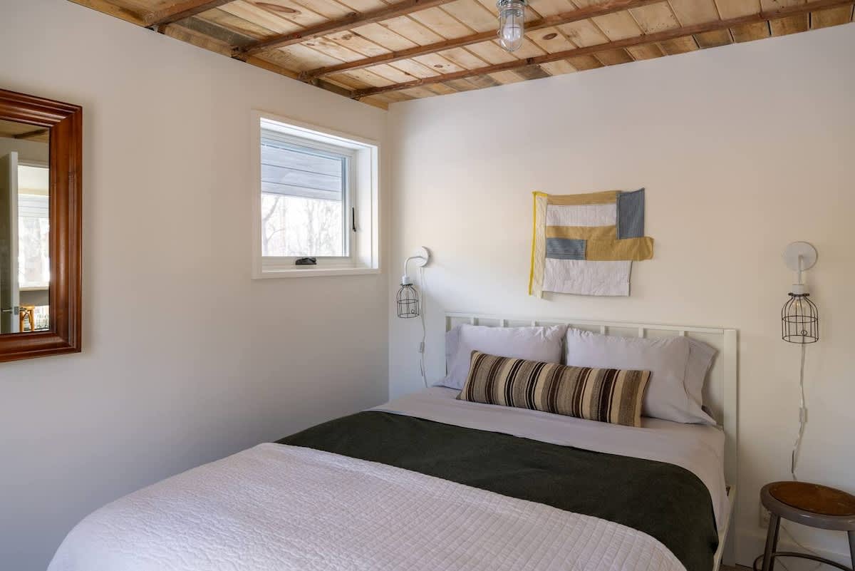 A cozy bedroom with a white metal-frame queen bed, light gray bedding, and a striped accent pillow, beneath a rustic wooden plank ceiling. A small window offers a glimpse of the outdoors.