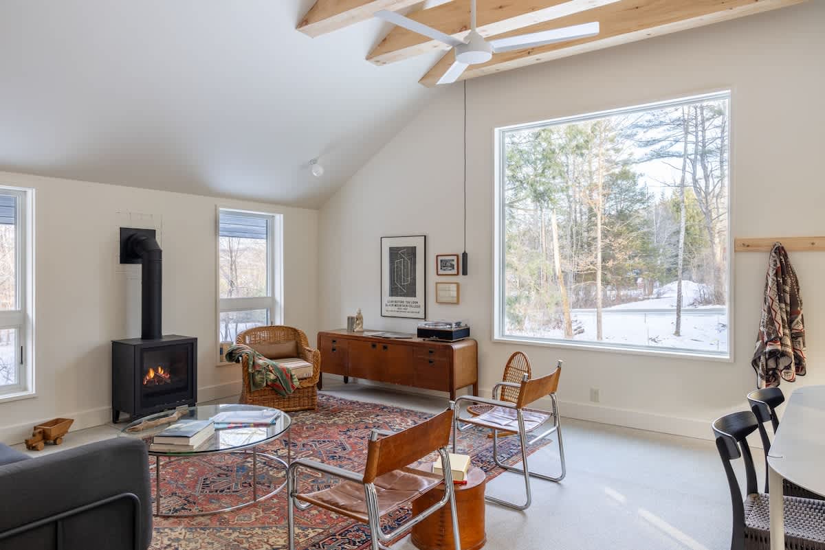 A spacious living room with a vaulted ceiling and exposed wooden beams, featuring a dark wood-burning stove, a wicker armchair, and a large window overlooking a snowy outdoor scene. A wooden credenza with a record player is visible.