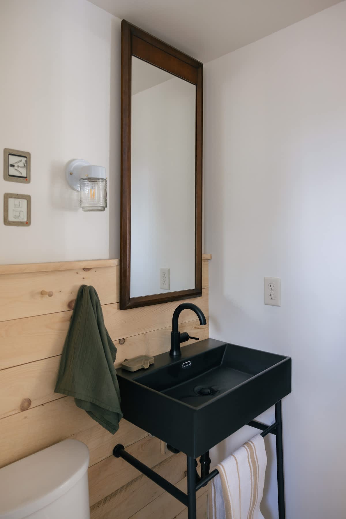 A modern bathroom features a black rectangular sink with a black faucet, set against wooden wainscoting and white walls, with a tall wooden-framed mirror above.