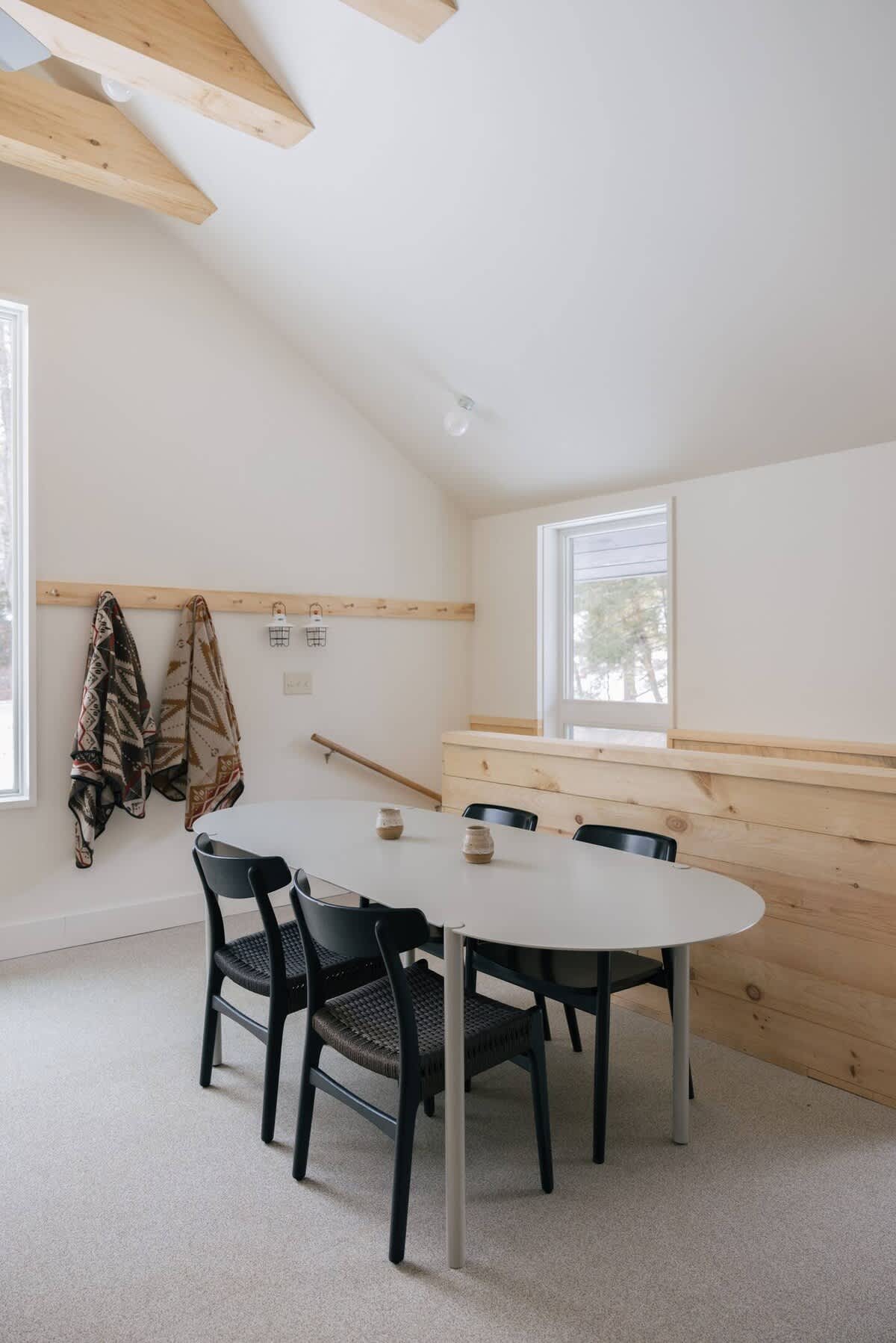 A bright dining area with a white oval table and four black chairs with woven seats, set on a light carpet. The room features a vaulted ceiling with exposed light wood beams and a wooden half-wall, with windows offering natural light.