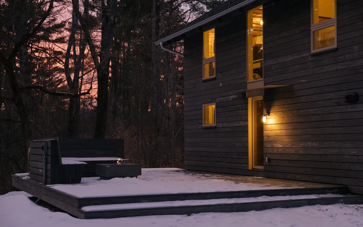 The dark grey modern house is bathed in the warm glow of a sunset, with interior lights on, highlighting the snowy deck and surrounding bare trees against an orange and pink sky.
